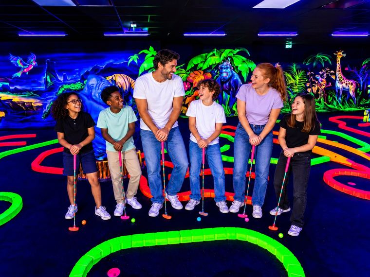 Deux parents et quatre enfants, club en main, devant les décors fluorescents du mini-golf Goolfy